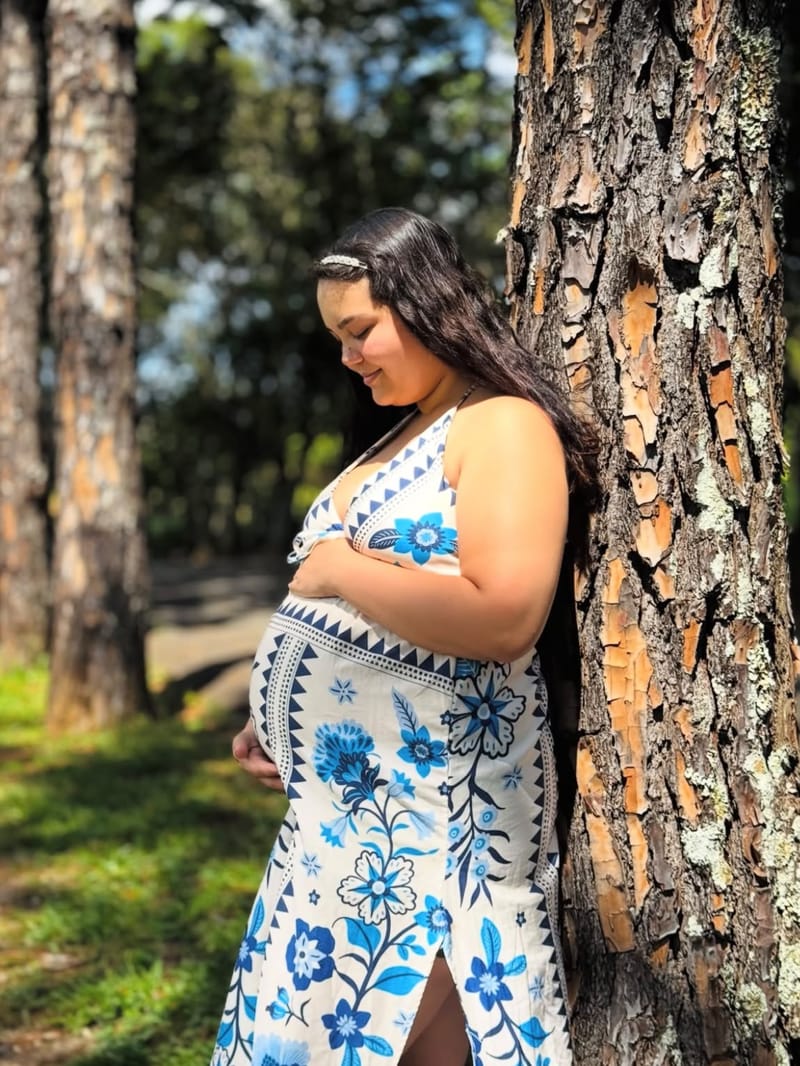 Gestante de vestido floral azul encostada em uma árvore, olhando para a barriga