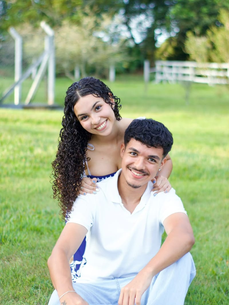 Casal sorrindo sentado na grama, ela abraçando ele por trás